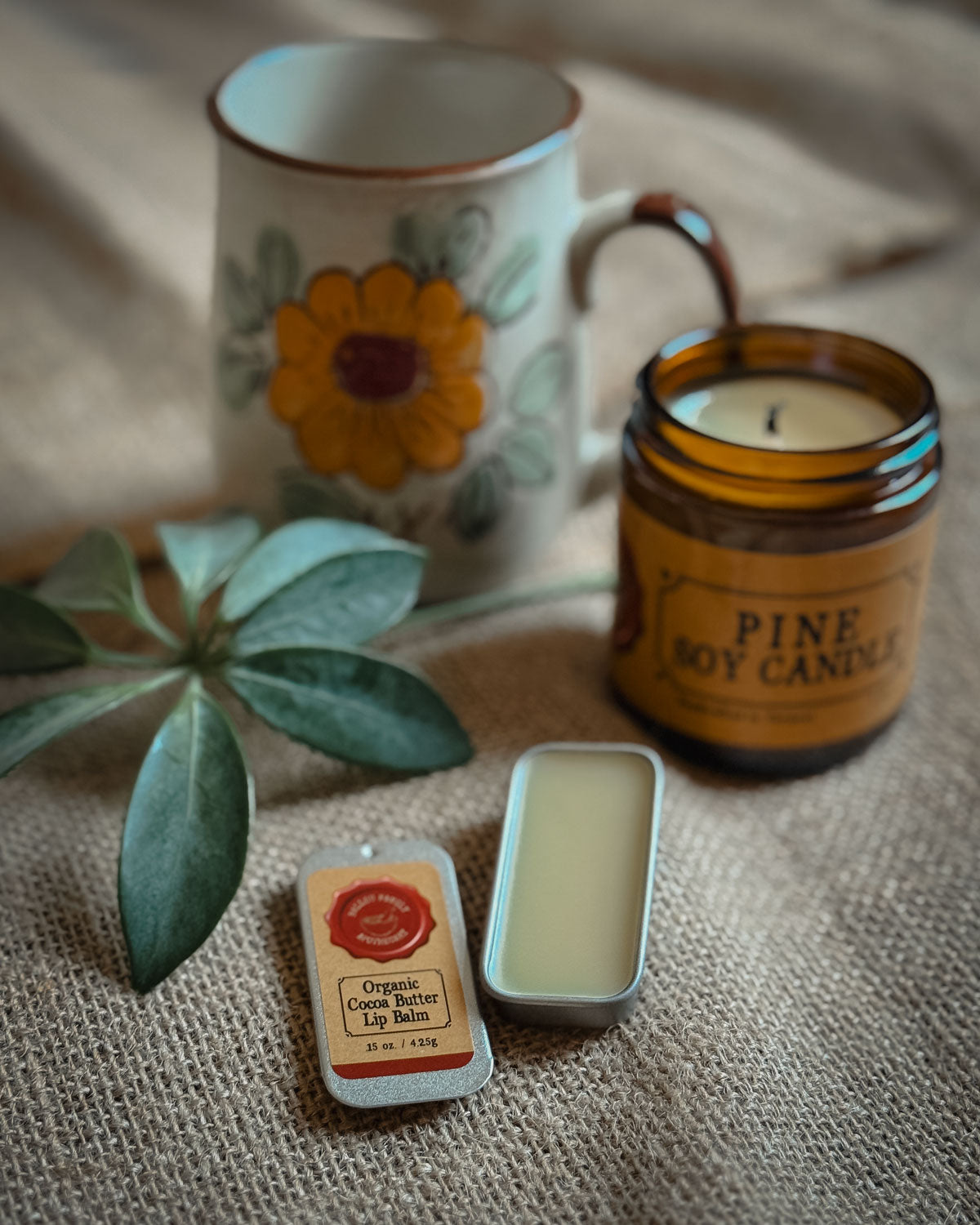 Tin of organic lip balm in front of a flower coffee mug and a soy candle