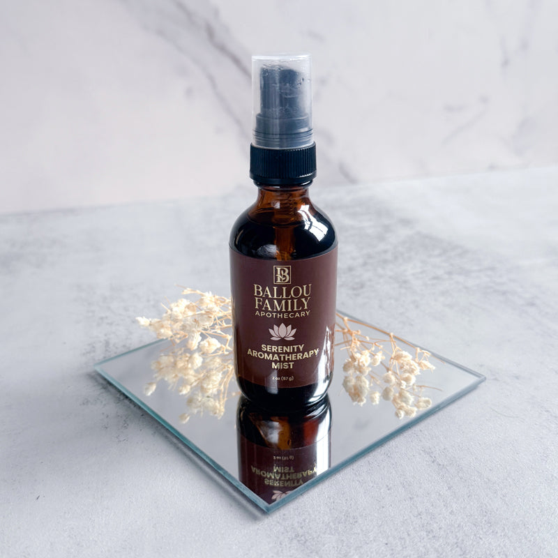 Bottle of Ballou Family Aromatherapy Serenity Mist on a reflective surface with flowers.