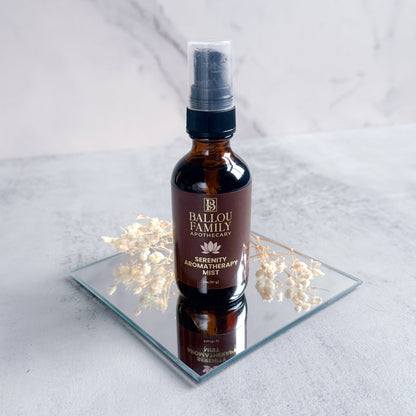 Bottle of Ballou Family Aromatherapy Serenity Mist on a reflective surface with flowers.