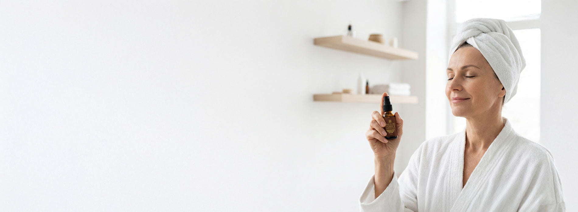 Woman in a white robe and towel holding a bottle of elixir in a bright room.