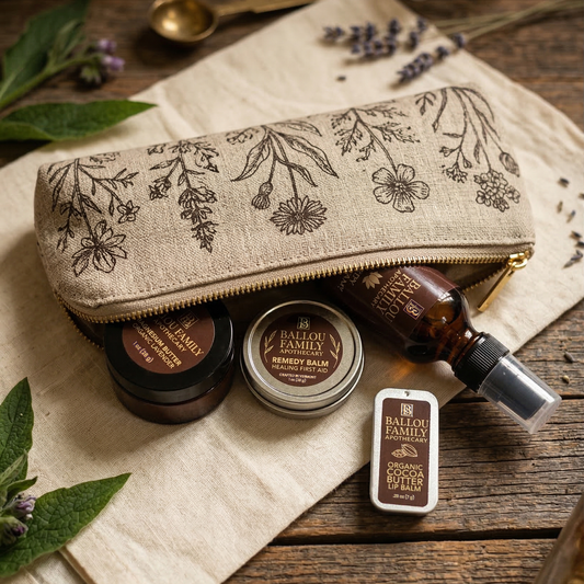 Ballou Family Apothecary products including a pouch, balm, and bottle on a wooden surface with plants.