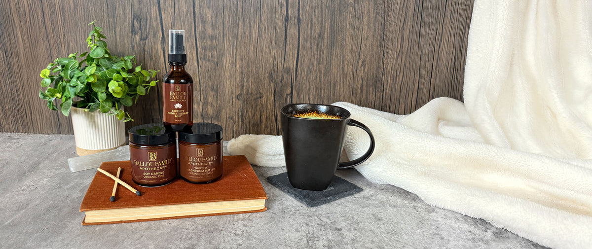 Three Ballou Family Apothecary Products on a book with a plant, mug and blanket