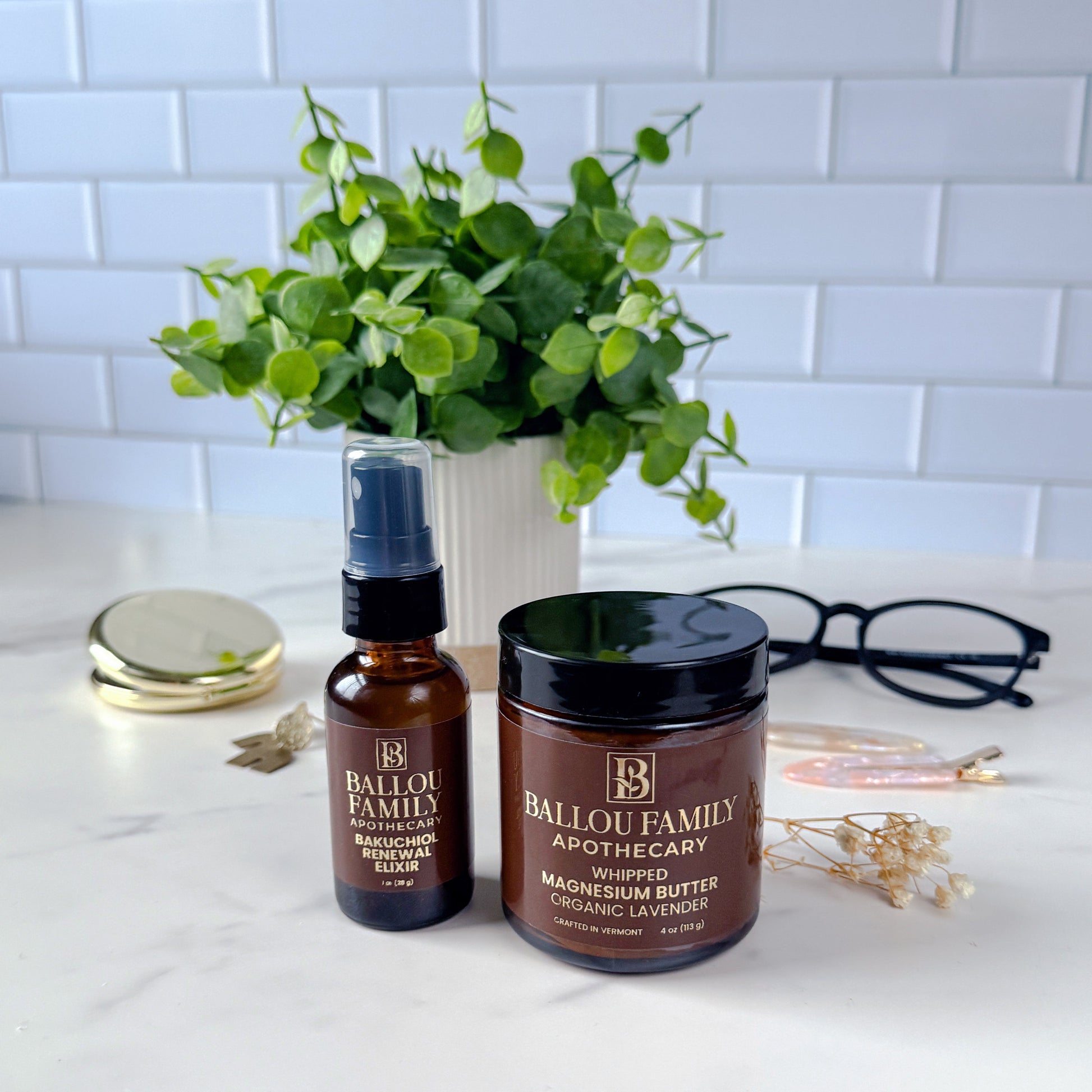 Two skincare products from Ballo Family Apothecary on a white surface with a plant and glasses in the background.