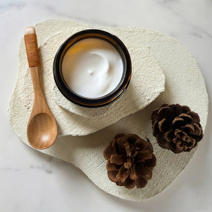 Jar of Magnesium Butter with a wooden spoon and pinecones on a textured stone surface.