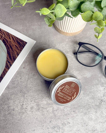 Open tin of Ballou Family Apothecary Remedy Balm with a plant and glasses in the background on a gray surface