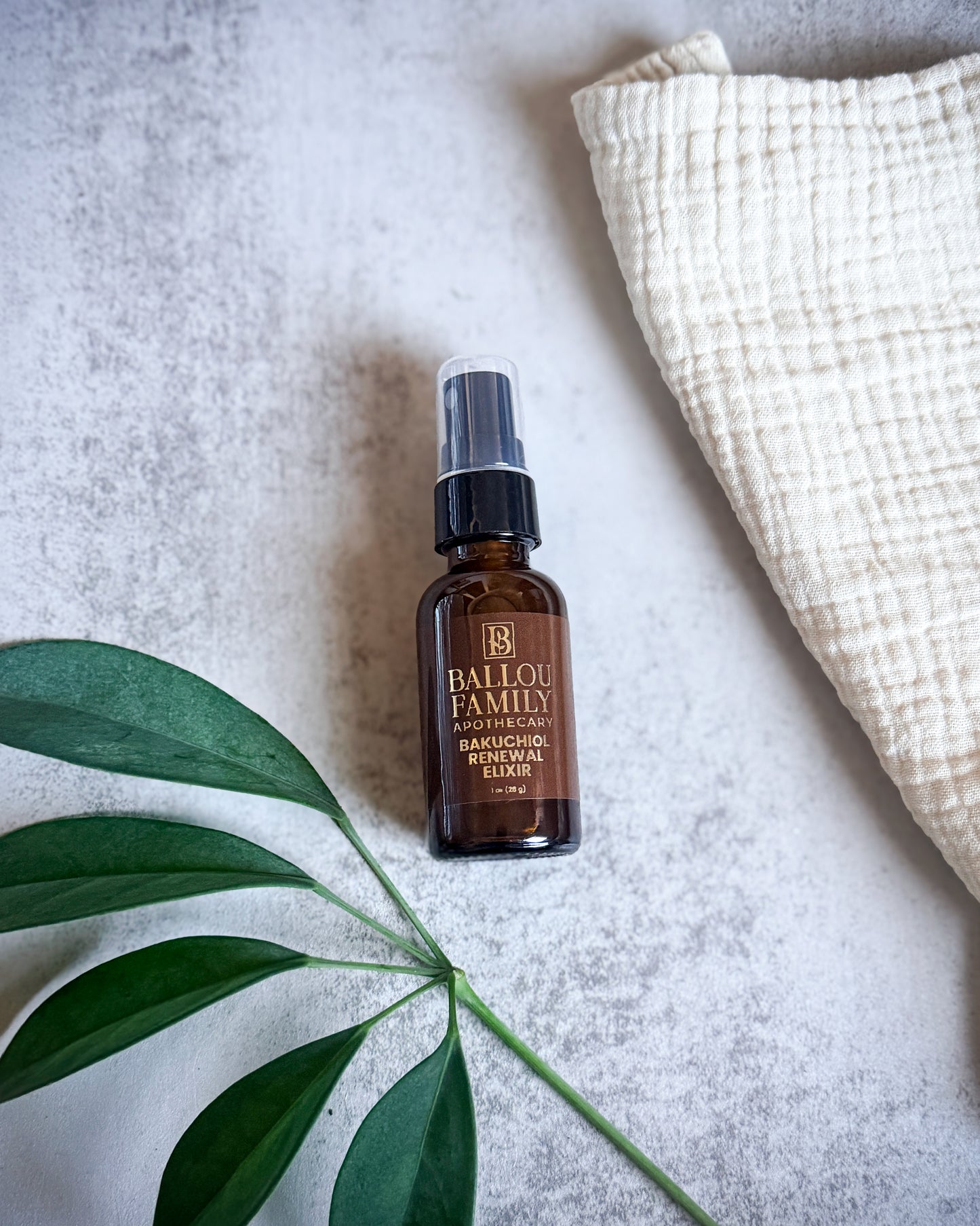 Brown bottle with black cap labeled 'Ballou Family Apothecary Bakuchiol Renewal Elixir' on a light gray surface with a green leaf and white fabric.
