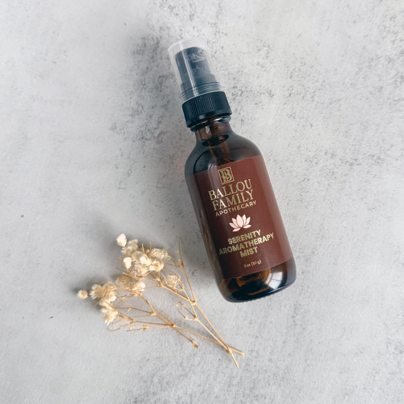 Brown spray bottle labeled 'Ballou Family Apothecary Serenity Aromatherapy Mist' on a gray surface with dried flowers.